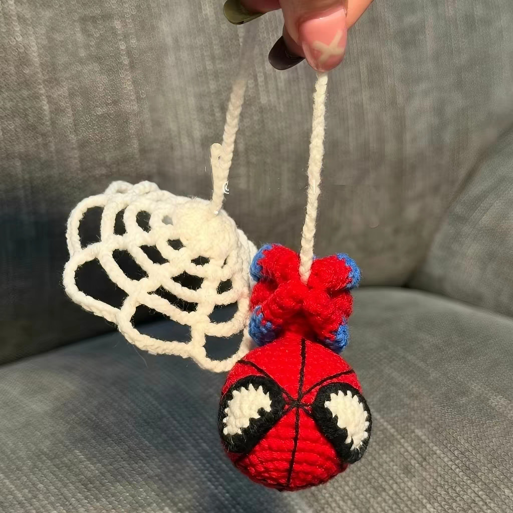 A small crocheted red-and-blue figure hanging upside down on a white chain thread beside a round white crocheted web, both held up from above over a grey sofa