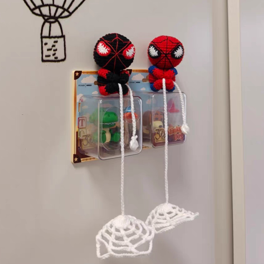 Two small crocheted figures, one black and red and one red and blue, sitting on top of two small boxed toys fixed to a wall, each with a white chain thread hanging down to a round white crocheted web