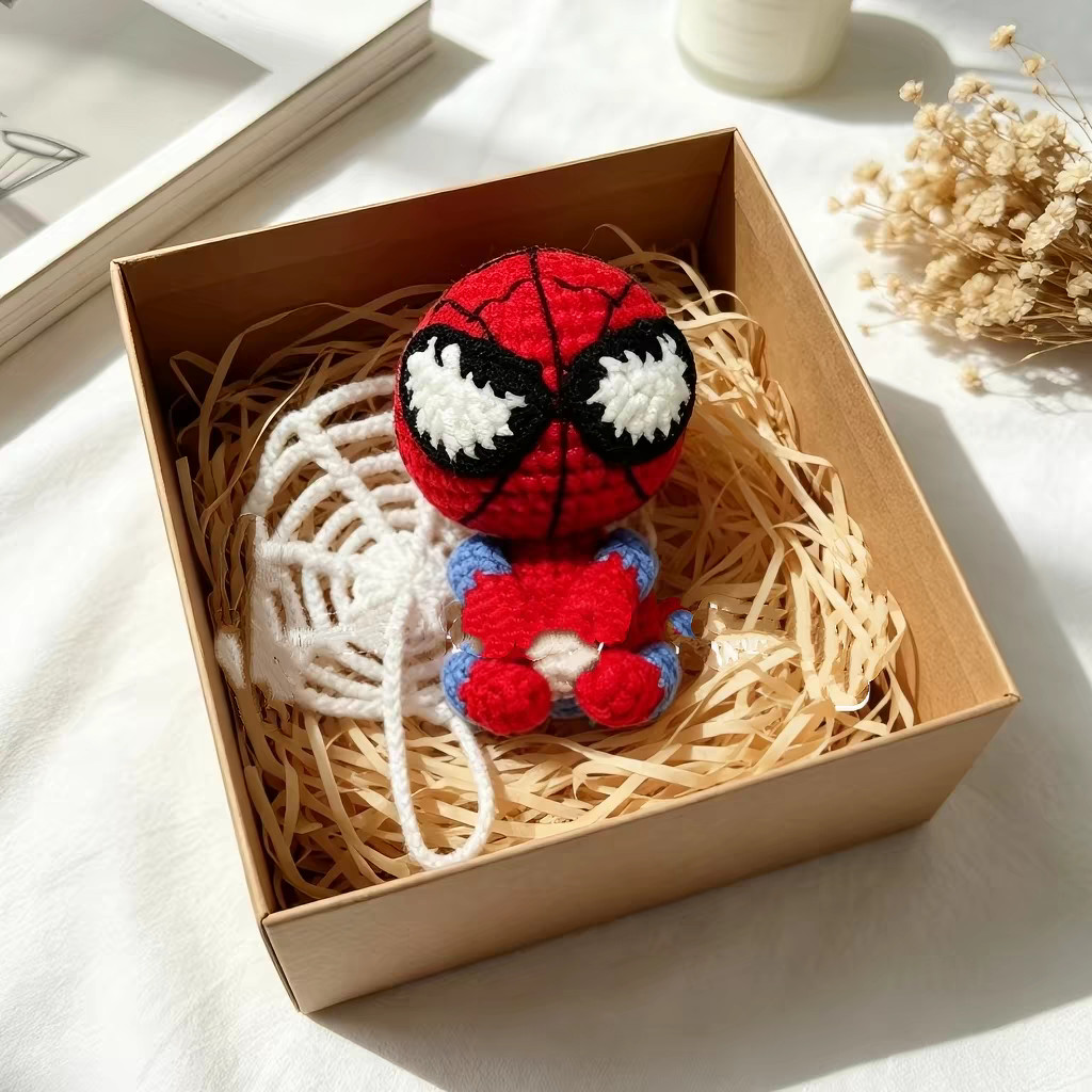 A small crocheted red-and-blue figure sitting on shredded paper in an open kraft box, a round white crocheted web and its chain thread beside it, with dried flowers and a book on a white cloth around the box
