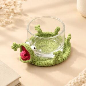 A crocheted sage green lizard coaster on a cream surface with a clear glass of water standing on it, the lizard's head at the near edge with its jaws open on a dark red mouth and a coiled pink tongue, two white eyes with black bead pupils above, and three-toed limbs and a short pointed tail around the corded rim of the disc