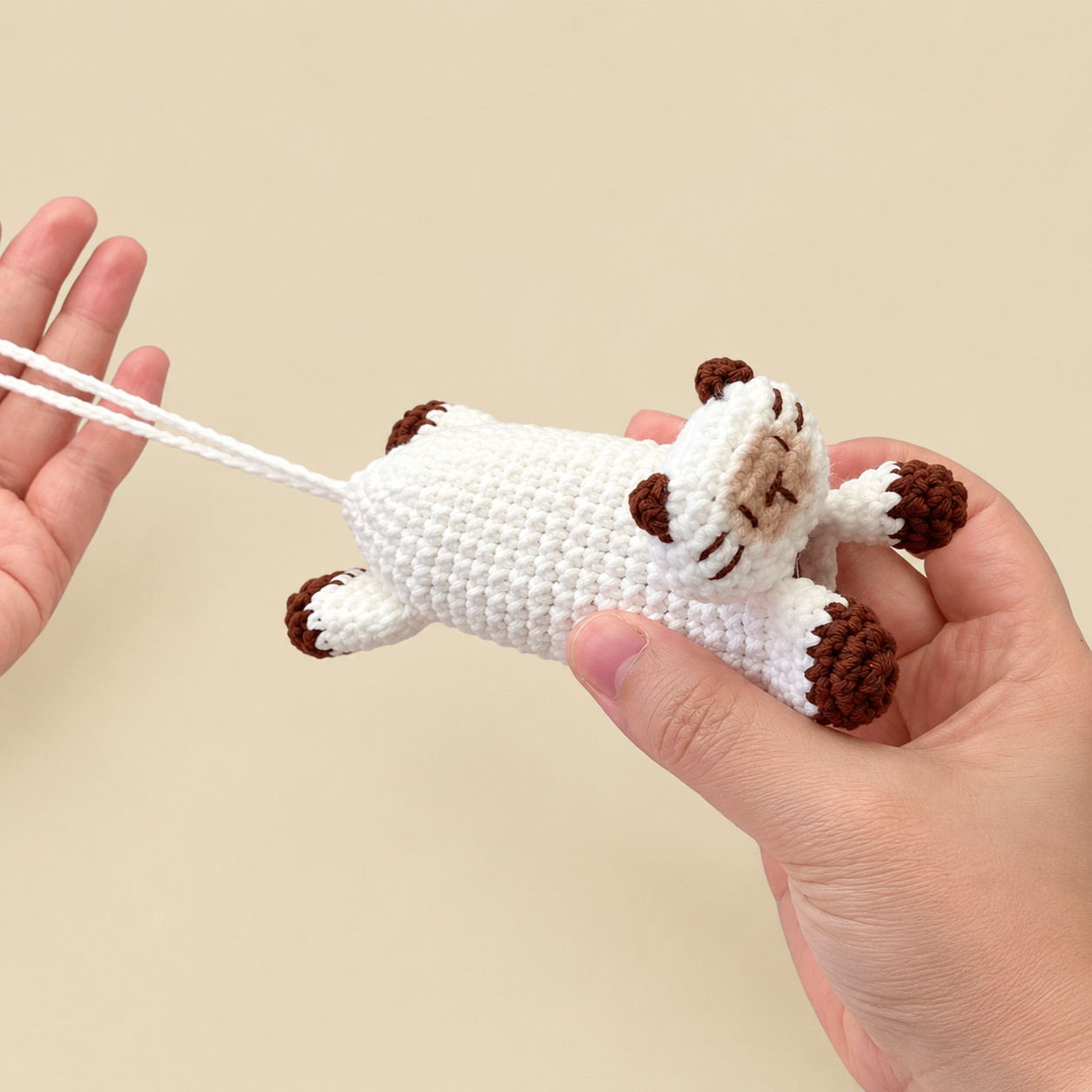 A white crocheted Siamese cat key pouch lying in an open hand against a beige background: chestnut ears, chestnut-tipped paws and legs, a beige muzzle with stitched chestnut eyes, nose and whiskers, and a long white chain loop running from its back end across the other open hand