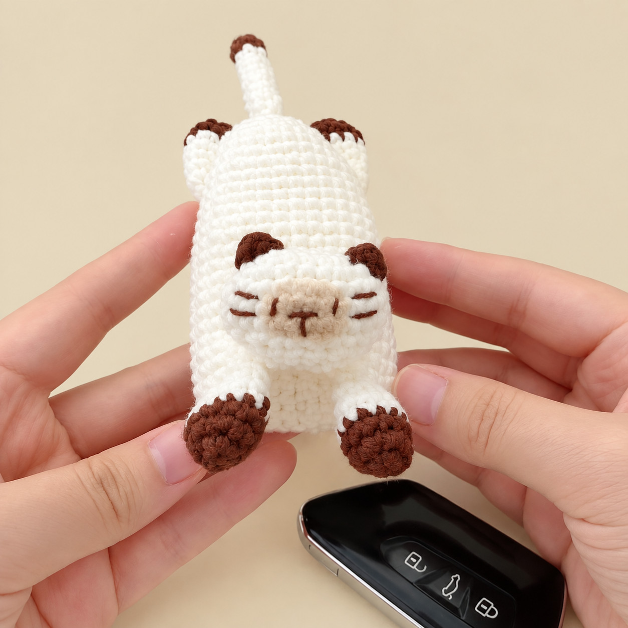A white crocheted Siamese cat key pouch held in two hands and seen from the front: a beige muzzle with stitched chestnut eyes, nose and whiskers, chestnut ears, two chestnut-tipped front paws, the hind legs and a chestnut-tipped tail behind, and a black car key fob below