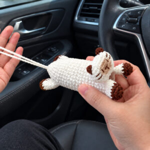 A white crocheted Siamese cat key pouch held in one hand inside a car, in front of the steering wheel: the cat lies stretched out along the pouch with chestnut ears, chestnut-tipped paws and legs, and a beige muzzle with stitched chestnut eyes, nose and whiskers, and a long white chain loop runs from its back end across the other open hand