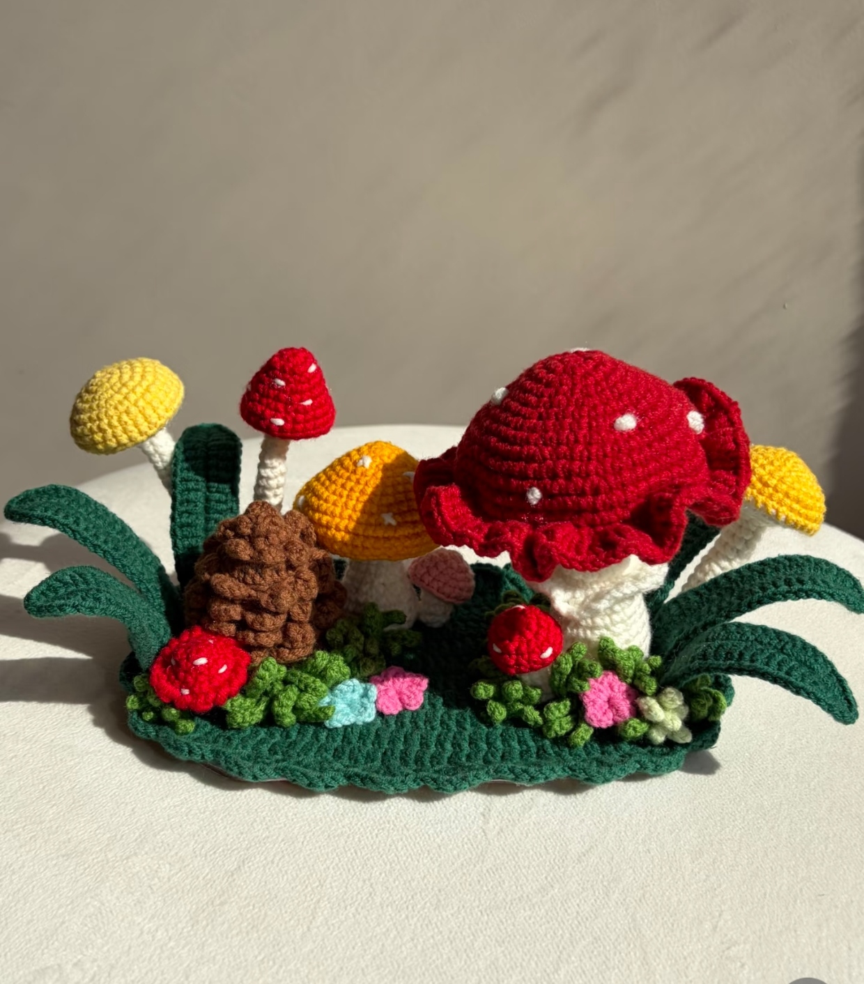 The same crocheted mushroom forest from the front on a cream cloth: the big red spotted toadstool, a brown pinecone, red, yellow and orange mushrooms on white stems, a small pink one, dark green leaves curling out at both ends, and grass and flowers along the scalloped green base