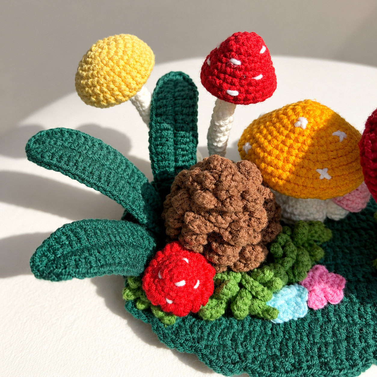 Close-up of the left end of the crocheted mushroom forest: a round yellow cap and a red cap with white dots on tall white stems, an orange cap with small white crosses, a brown pinecone of rounded scales, a small red mushroom with white dots, curly green grass, a blue and a pink flower, and dark green leaves