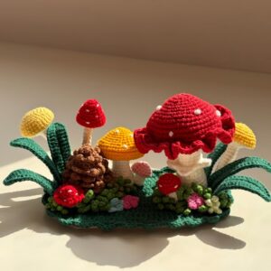 A crocheted mushroom forest on a green scalloped base in sunlight: a big red toadstool with white dots and a ruffled rim on a white stem, a brown pinecone, smaller mushrooms with red, yellow, orange and pink caps, long dark green leaves, curly green grass and small pink, blue and cream flowers