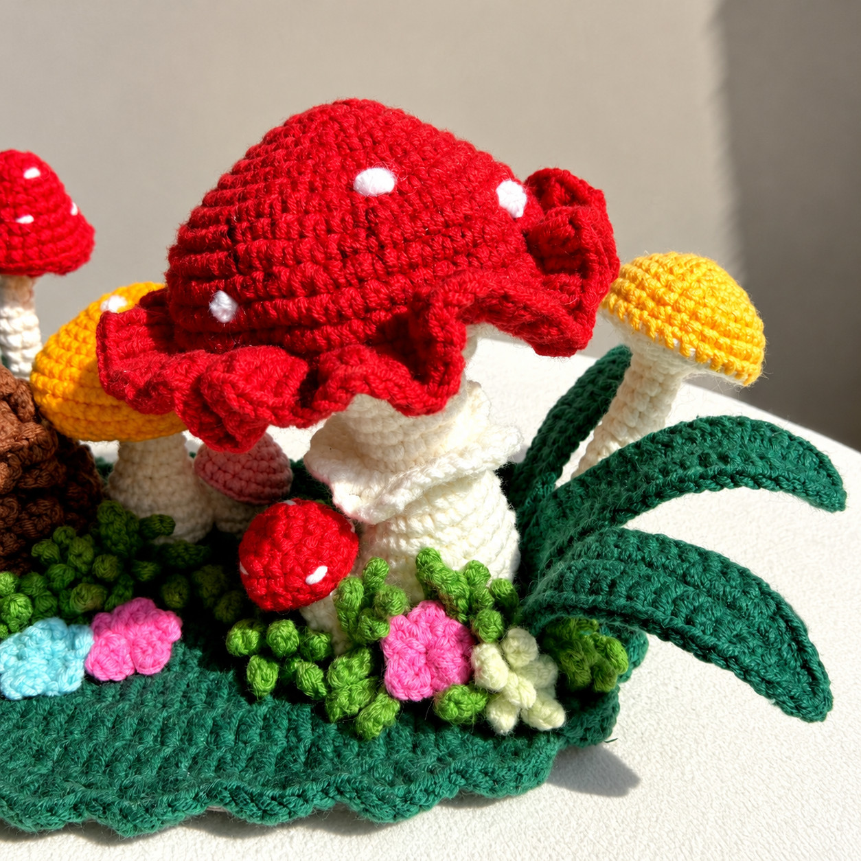 Close-up of the big crocheted toadstool: a red cap with white dots and a deep ruffled rim over a white stem with a frill, a small red mushroom with white dots in front of it, a yellow mushroom and curling dark green leaves to the right, and green grass with pink, cream and blue flowers along the base
