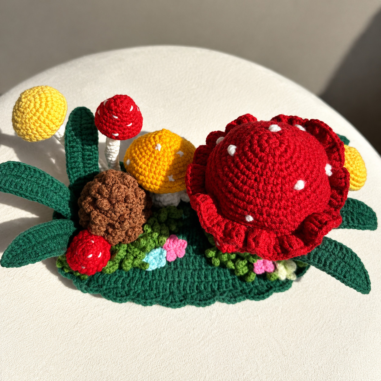 The crocheted mushroom forest seen from above on a round cream stool: the big red toadstool's dotted cap and ruffled rim, a brown pinecone, red, yellow and orange mushroom caps, long dark green leaves spreading out from both ends of the oval base, and grass with small pink, blue and cream flowers