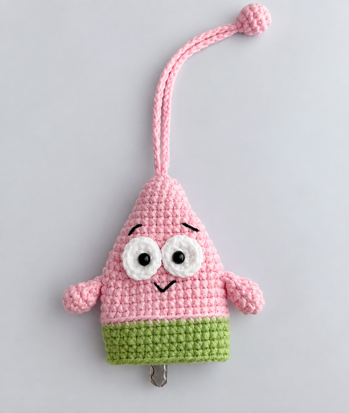 A crocheted pink starfish key pouch seen straight on against a pale grey background: white eyes with black pupils, stitched black eyebrows and a small smile, an arm on each side, green shorts below a ridge where the pink ends, a long pink cord looped up to a small ball, and the tip of a key showing below