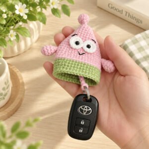 A crocheted pink starfish key pouch held in an open hand: white eyes with black pupils, stitched black eyebrows and a small smile, two stubby arms and green shorts, a small pink ball on top, and a black car key fob on a metal ring drawn out on the cord below