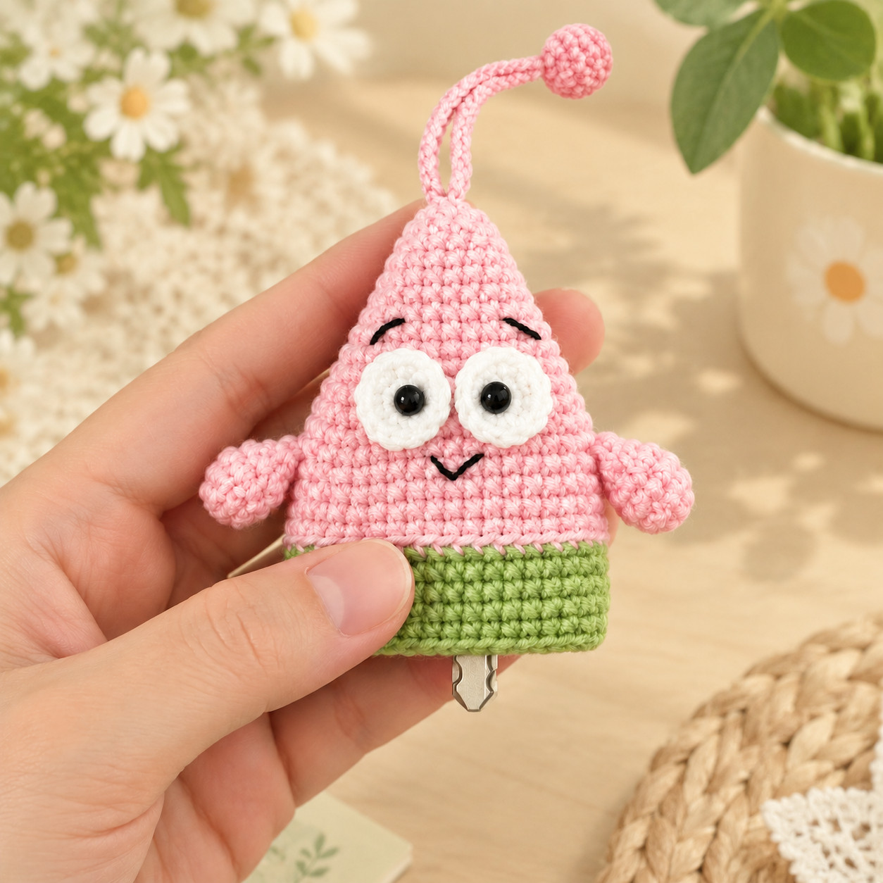 A crocheted pink starfish key pouch held in a hand, face on: white eyes with black pupils, stitched black eyebrows and a small smile, two stubby arms and green shorts, a pink cord looped up to a small ball, and the tip of a key poking out below