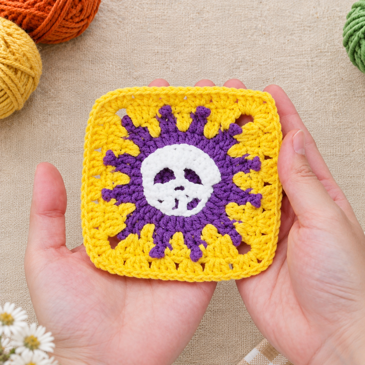 Two hands holding a yellow granny square with a purple sunburst and a white crocheted skull in the middle, on cream linen beside balls of yarn