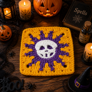 A yellow granny square with a purple sunburst and a white crocheted skull, on dark wood beside a lit jack-o'-lantern, candles, a book marked Spells, a Witch's Brew bottle and a witch's hat