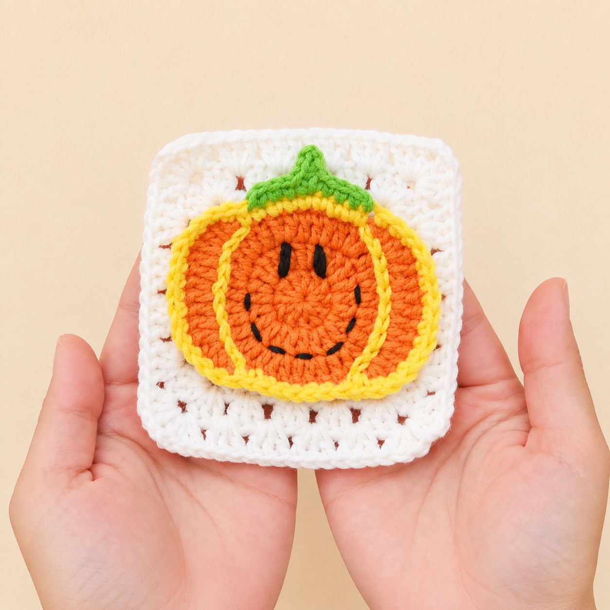 Two hands holding a white granny square with an orange crocheted pumpkin on it, against a plain peach background