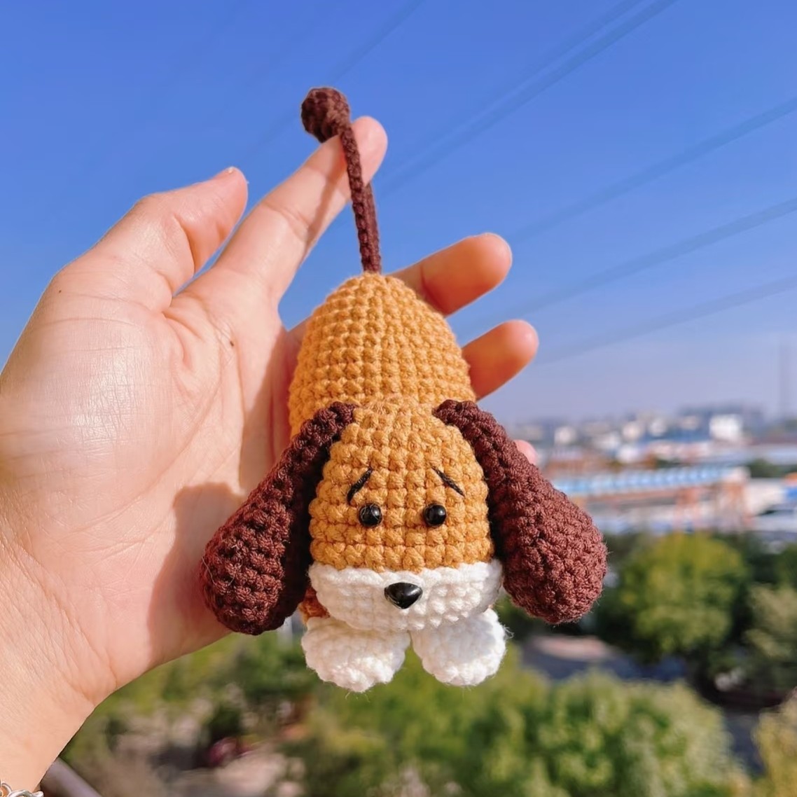 A crocheted puppy key pouch held up against a blue sky: a golden-brown body with a dark brown tail curling up from the closed end, a head with black eyes and stitched eyebrows, a black nose on a white muzzle, dark brown floppy ears and two white paws