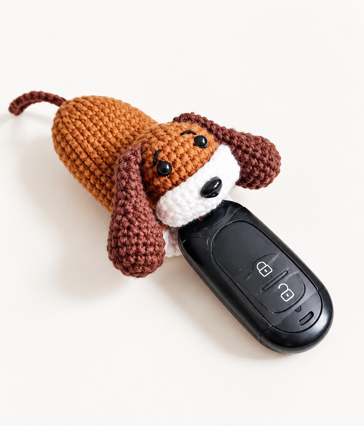 A crocheted puppy key pouch lying on a white surface: a ginger-brown body with a dark brown tail, black eyes, stitched eyebrows and a black nose on a white muzzle, dark brown floppy ears, and a black car key fob half drawn out of the opening
