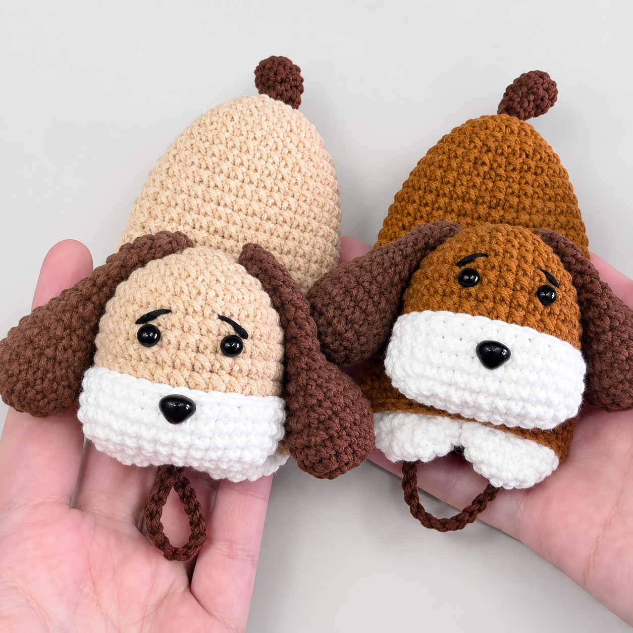 Two crocheted puppy key pouches held side by side in two hands, one beige and one ginger-brown, each with dark brown floppy ears, a round tail at the top, black eyes, stitched eyebrows, a black nose on a white muzzle and a brown chain loop hanging below