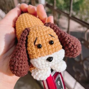 A crocheted puppy key pouch held in an open hand in sunlight: a golden-brown head with black eyes and stitched eyebrows, a black nose on a white muzzle, dark brown floppy ears and two white paws, with a red car key fob drawn out from under the paws