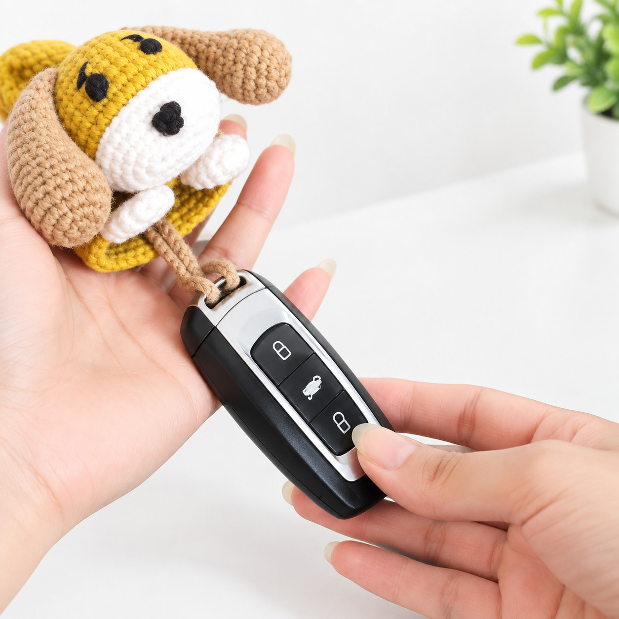 A mustard crocheted puppy key pouch with tan floppy ears, black eyes, a black nose on a white muzzle and two white paws, resting in one hand, with a black car key fob hanging from its tan chain loop and held in the other hand
