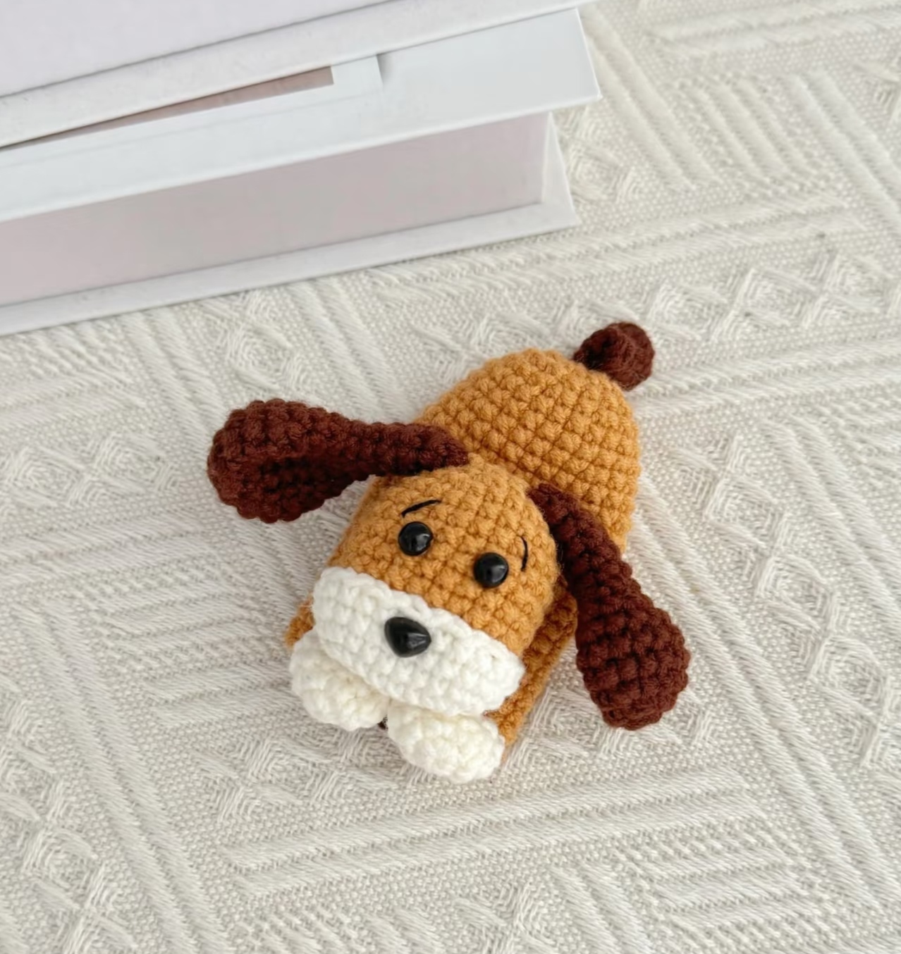 A crocheted puppy key pouch lying on a cream textured blanket beside a white box: a golden-brown body with a dark brown tail, black eyes, stitched eyebrows, a black nose on a white muzzle, dark brown floppy ears and two white paws