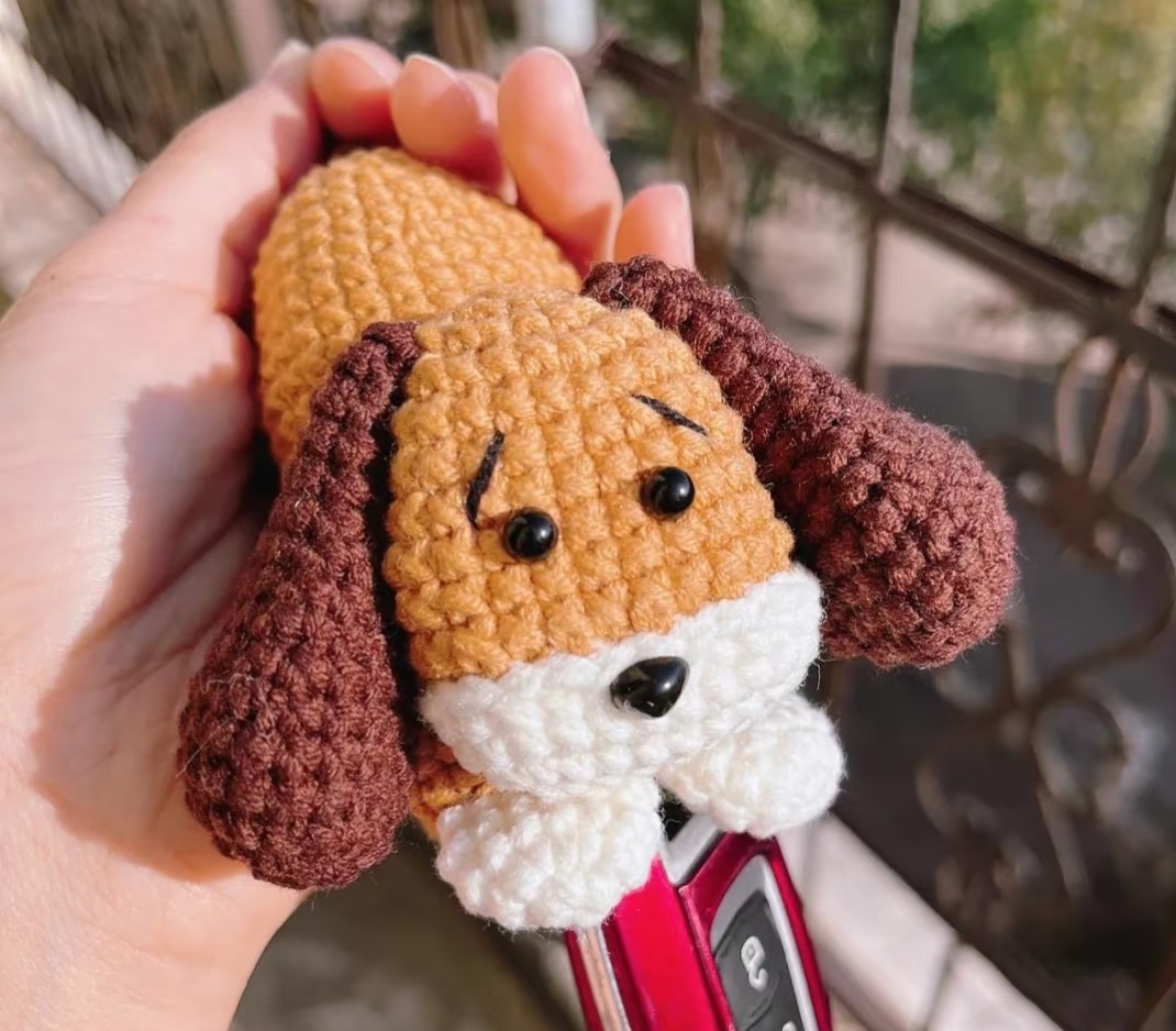 A crocheted puppy key pouch held in an open hand in sunlight: a golden-brown head with black eyes and stitched eyebrows, a black nose on a white muzzle, dark brown floppy ears and two white paws, with a red car key fob drawn out from under the paws