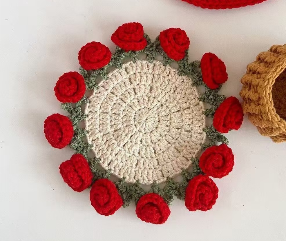 A rose coaster on a white surface: a cream centre worked in rounds, a sage green lacy edging with little leaf points, and red rolled roses round the rim, with the edge of a tan crocheted flower pot beside it