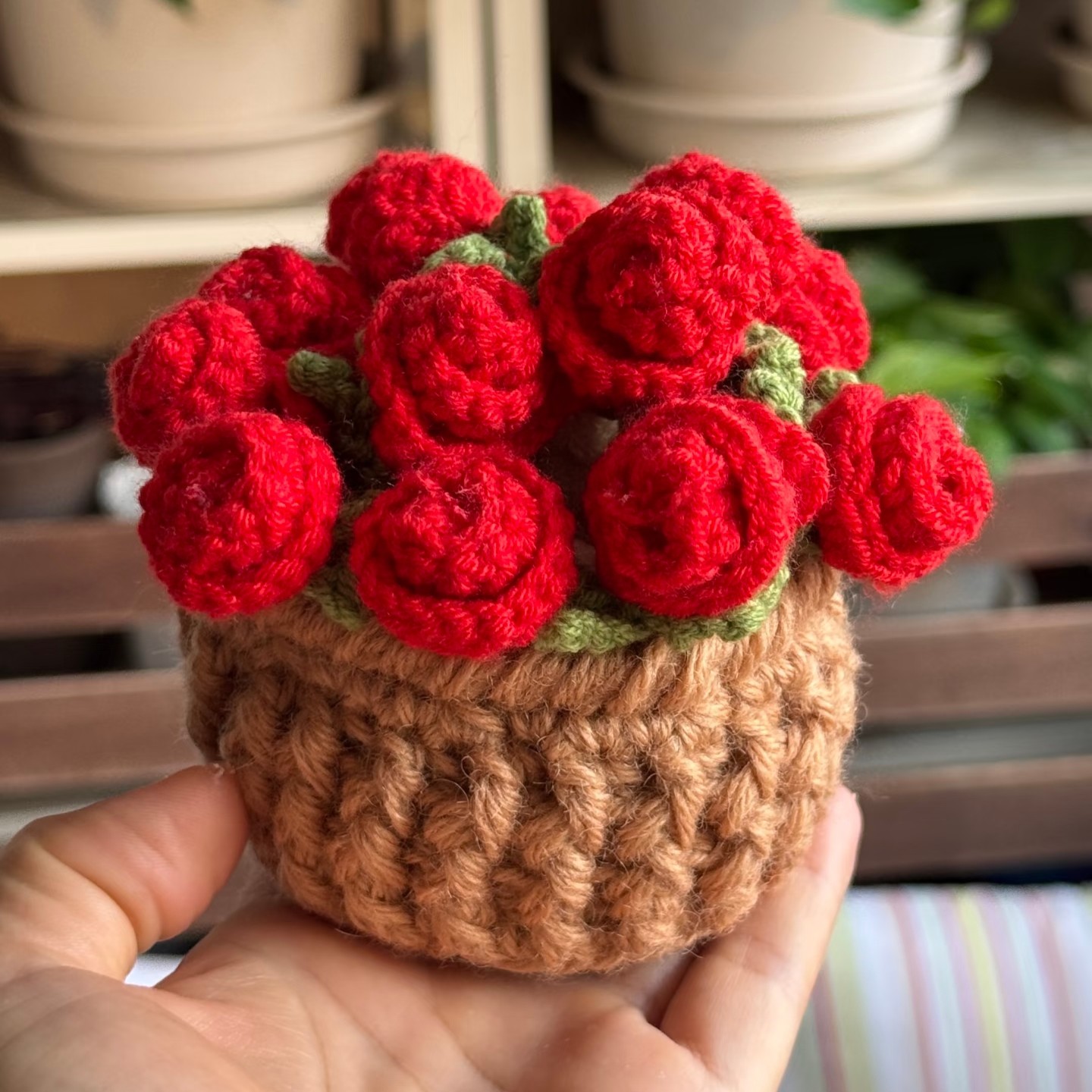 A small bunch of red crocheted roses with green leaves standing in a tan ribbed crocheted flower pot, held on an open hand in front of shelves of plant pots