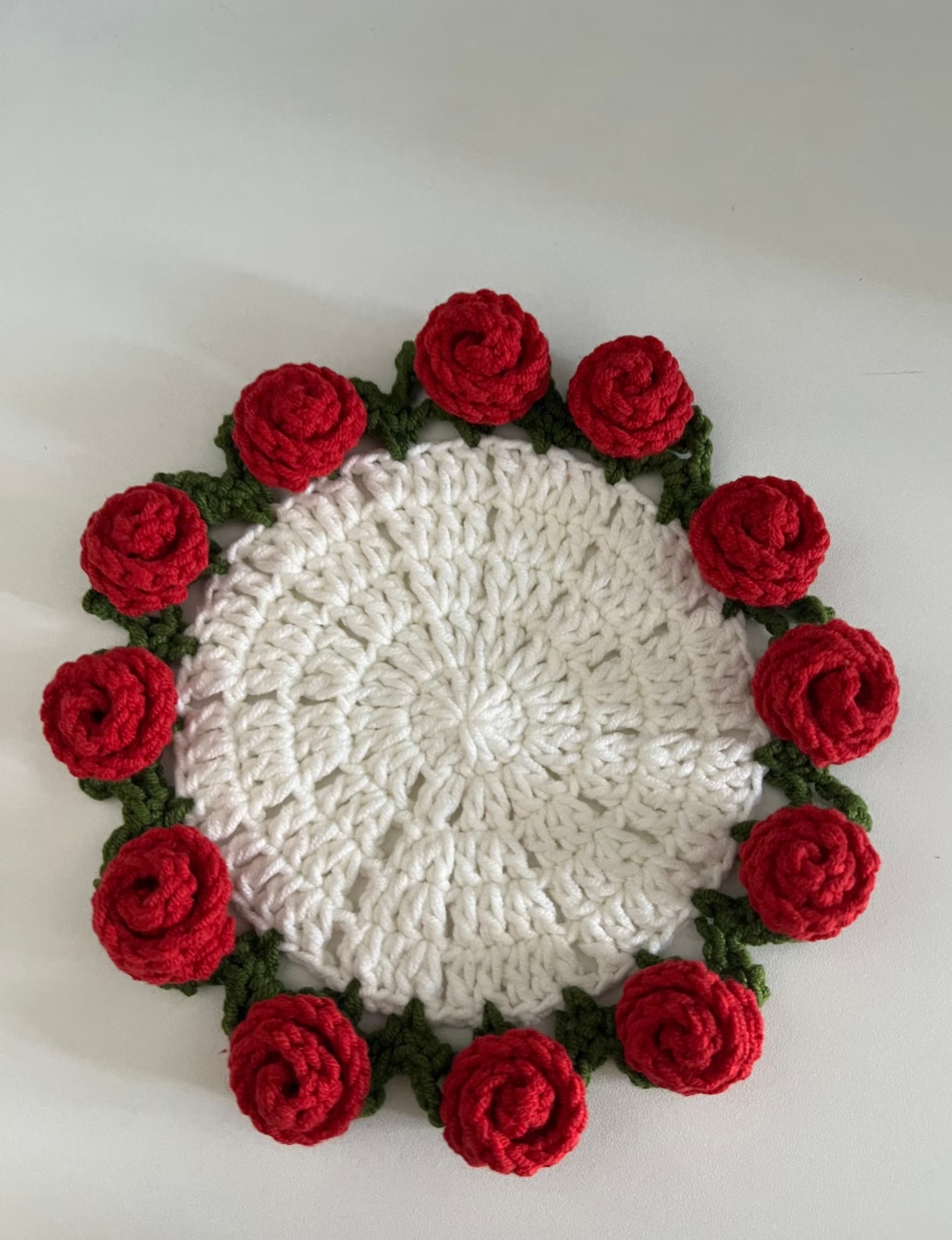 A rose coaster seen from above on a white surface: a bright white centre of double crochet rounds with small holes between the stitches, a dark green edging of leaf points and red rolled roses round the rim