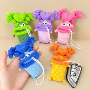 Five crocheted sausage-lip key pouches on a beige background, in green, lilac, pink, light blue and yellow, each with a thick rolled lip, small white eyes with black pupils, a heart on top between two tufted bunches of yarn hair, and a white cord hanging from its mouth; the blue one rests in an open hand, and the yellow one, with an orange lip, has its cord tied to a silver car key fob