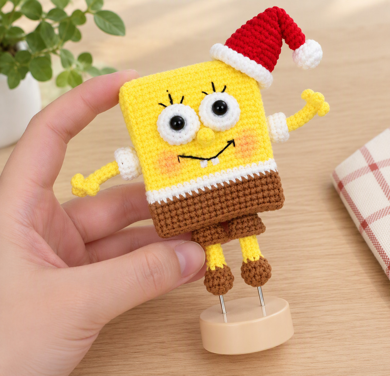 A crocheted yellow sea sponge in a red Santa hat held in one hand, its brown shoes resting on two wires in a round wooden stand: big white eyes with eyelashes, a small yellow nose, a black smile with two white teeth, white sleeves, a white shirt band and brown shorts