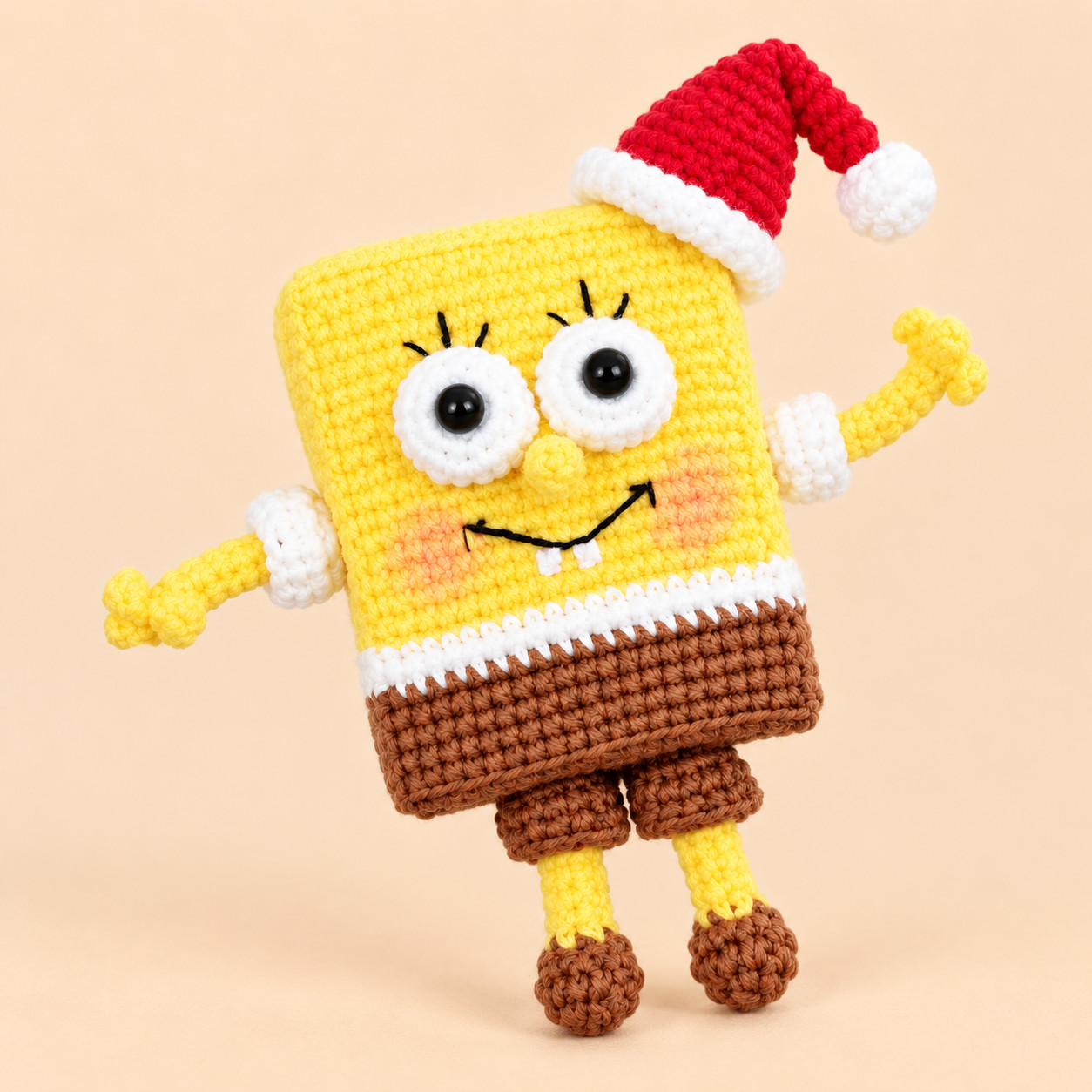 A crocheted yellow sea sponge standing face on against a peach background: a red Santa hat flopped to one side with a white brim and pompom, big white eyes with black centres and eyelashes, a small yellow nose, a black smile with two white teeth, pink cheeks, one arm raised, a white shirt band, brown shorts and thin yellow legs in brown shoes