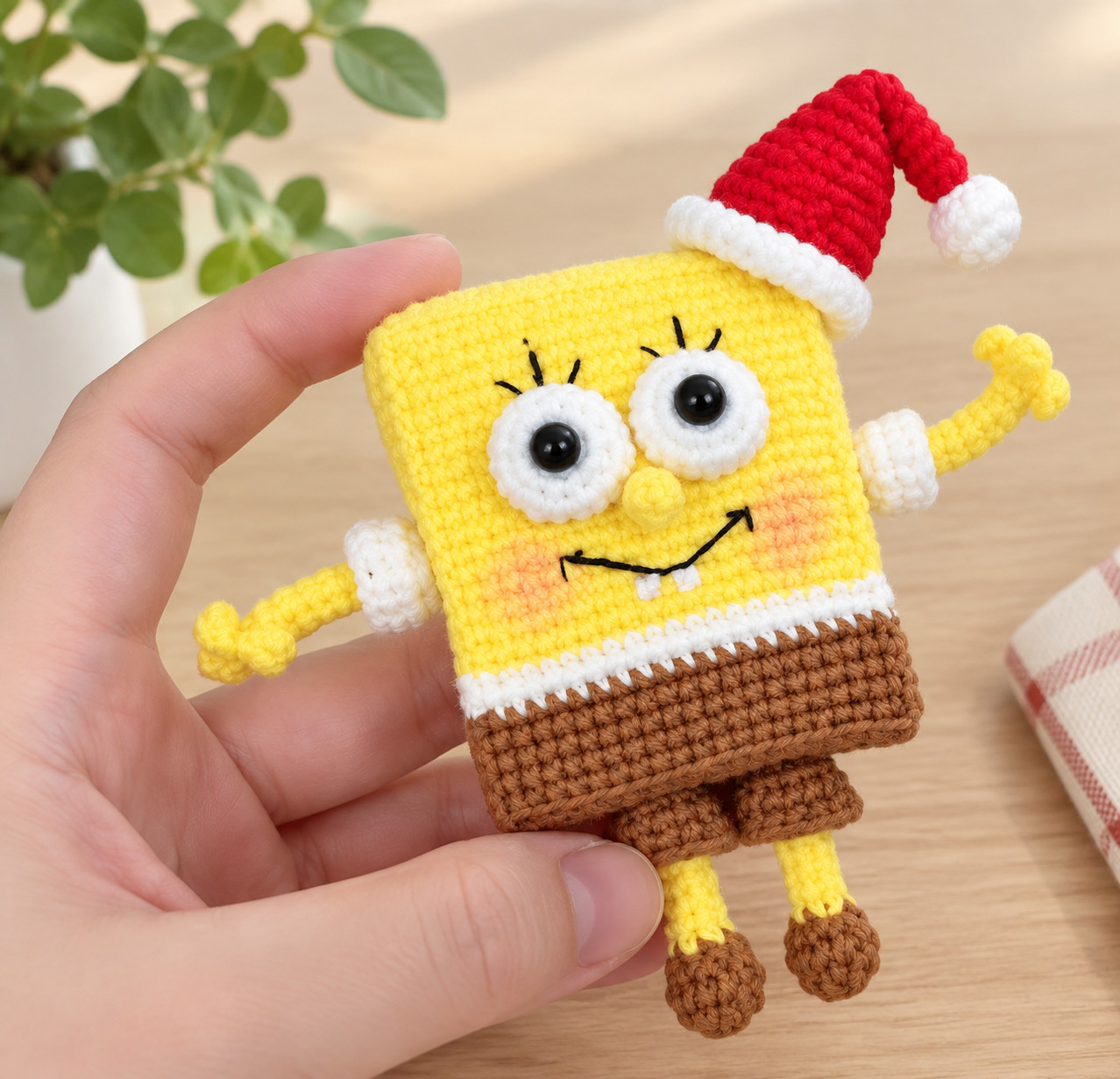 A crocheted yellow sea sponge held in one hand, face on: a red Santa hat with a white brim and pompom, big white eyes with black centres, a black smile with two white teeth, pink cheeks, three-fingered yellow arms in white sleeves, brown shorts and thin yellow legs in brown shoes