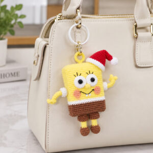 A crocheted yellow sea sponge hanging by a small yellow loop from a white ring and a gold clasp on a cream handbag: big white eyes with eyelashes, a wide black smile with two white teeth, pink cheeks, a red Santa hat with a white brim and pompom, thin yellow arms in white sleeves, brown shorts and brown shoes