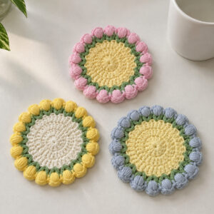 Three crocheted tulip coasters on a white table beside a white mug and a leafy plant: pink tulips round a yellow centre, yellow tulips round a white centre, and blue tulips round a yellow centre, each ring of tulips standing in a round of green V's