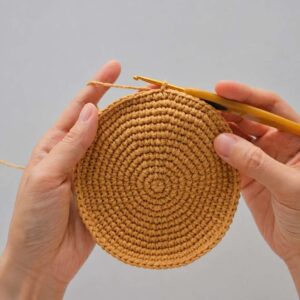 Two hands holding a flat, round caramel-coloured crochet disc worked in rounds of single crochet from a small centre, with a yellow-handled hook still in the edge and the working yarn running over one finger, against a plain grey background