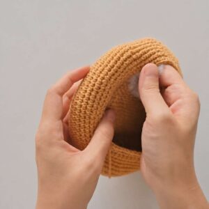 Two hands pressing the rim of the caramel-coloured crochet bowl, rolled over into a thick ring, with white stuffing showing inside the roll under the right thumb