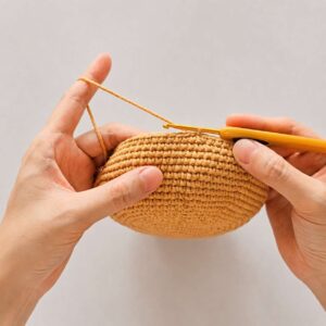 Two hands holding the caramel-coloured crochet bowl side-on, its wall curving up from a rounded base, with a yellow-handled hook in the top edge and the working yarn wrapped over the left forefinger