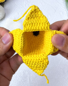 The yellow crocheted peel before its linings go on, held open in two hands: four flaps opened out round a square opening, a ridge of loose loops along the base of each, and the dark brown tip at the bottom of the cup