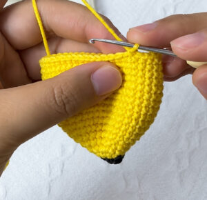 The yellow crocheted peel held in the fingers before its flaps, a silver crochet hook still in its top edge and the working yarn running up, with its dark brown tip at the bottom