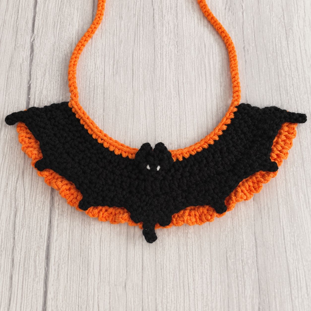 The bat collar laid flat on pale wood, showing the black wings, the four small points along the edge, the tail at the centre, the orange ruffle behind and the two orange ties