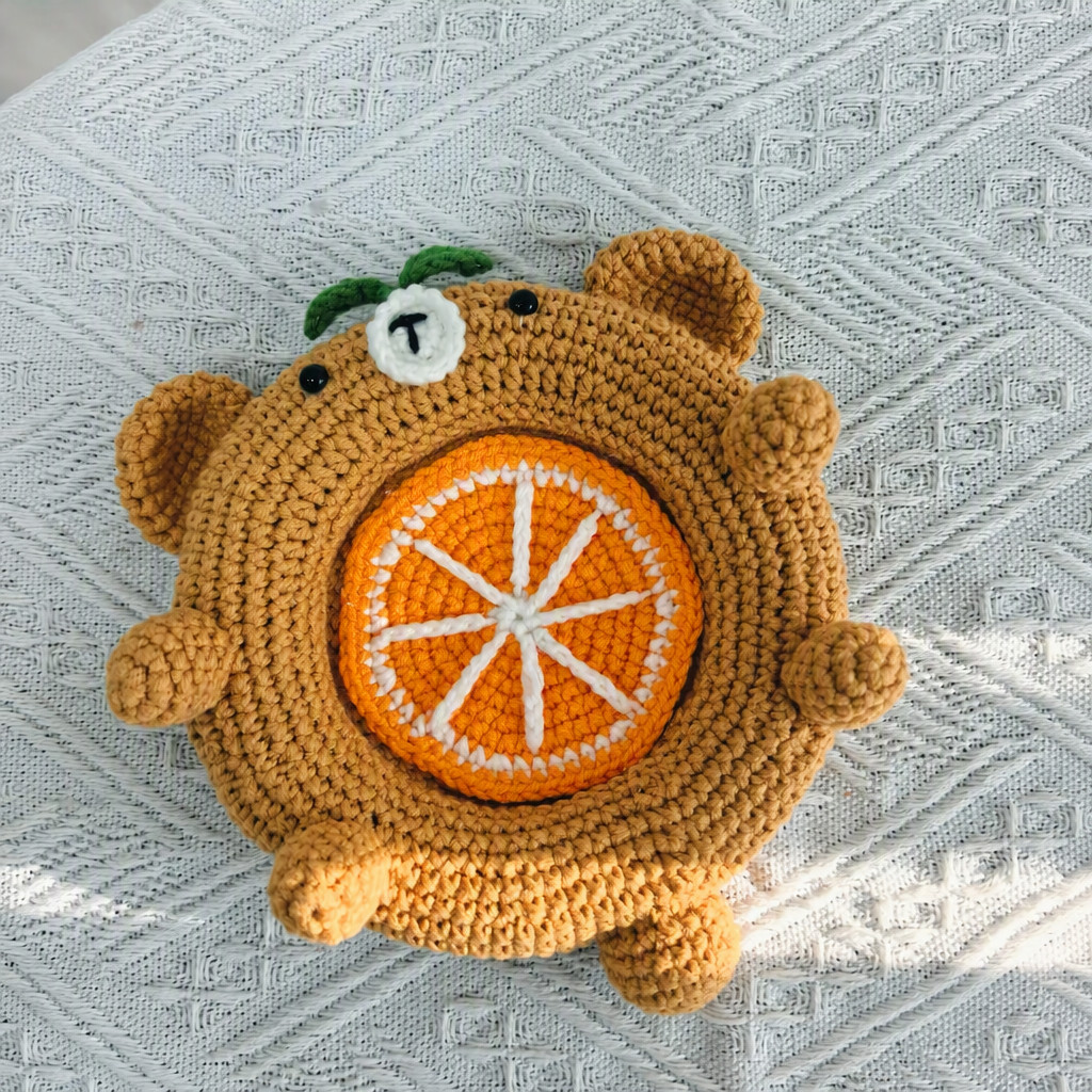 A caramel crocheted bear dish seen from above on a white textured blanket, with an orange slice sitting in the well, white segment lines running across it, two round ears, a white muzzle, two green leaves on its head and round paws around the padded rim