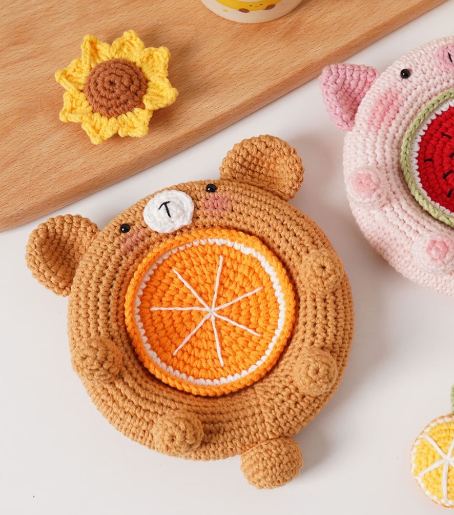 The caramel crocheted bear dish on a white table with an orange slice in the well, a white muzzle and pink cheeks, next to a crocheted sunflower on a wooden board and part of a pink pig dish with a watermelon slice