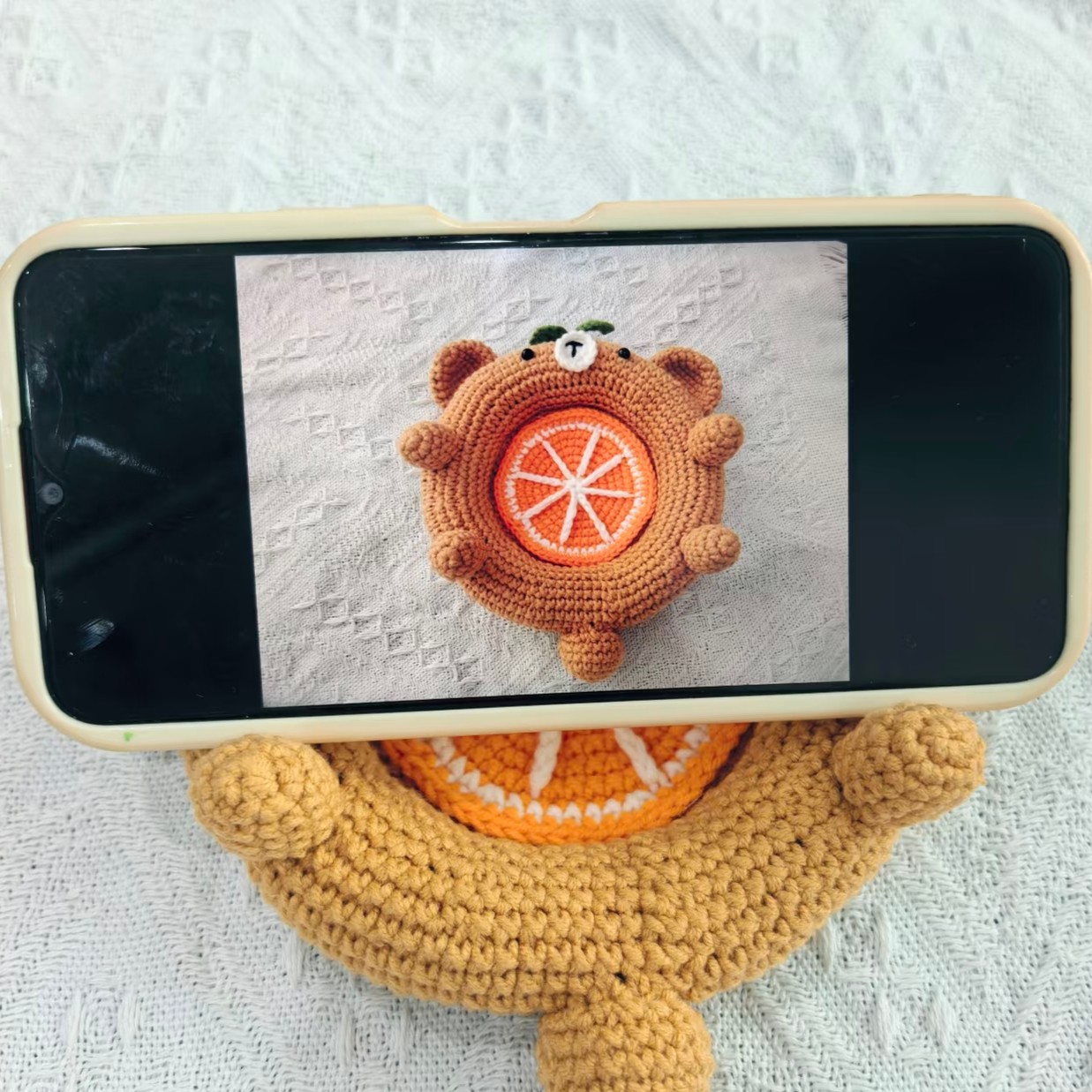 A phone lying sideways across the caramel crocheted bear dish on a white textured blanket, held up by the bear's two front paws, its screen showing a photo of a bear dish with an orange slice, and the real orange slice and the bear's tail showing below the phone