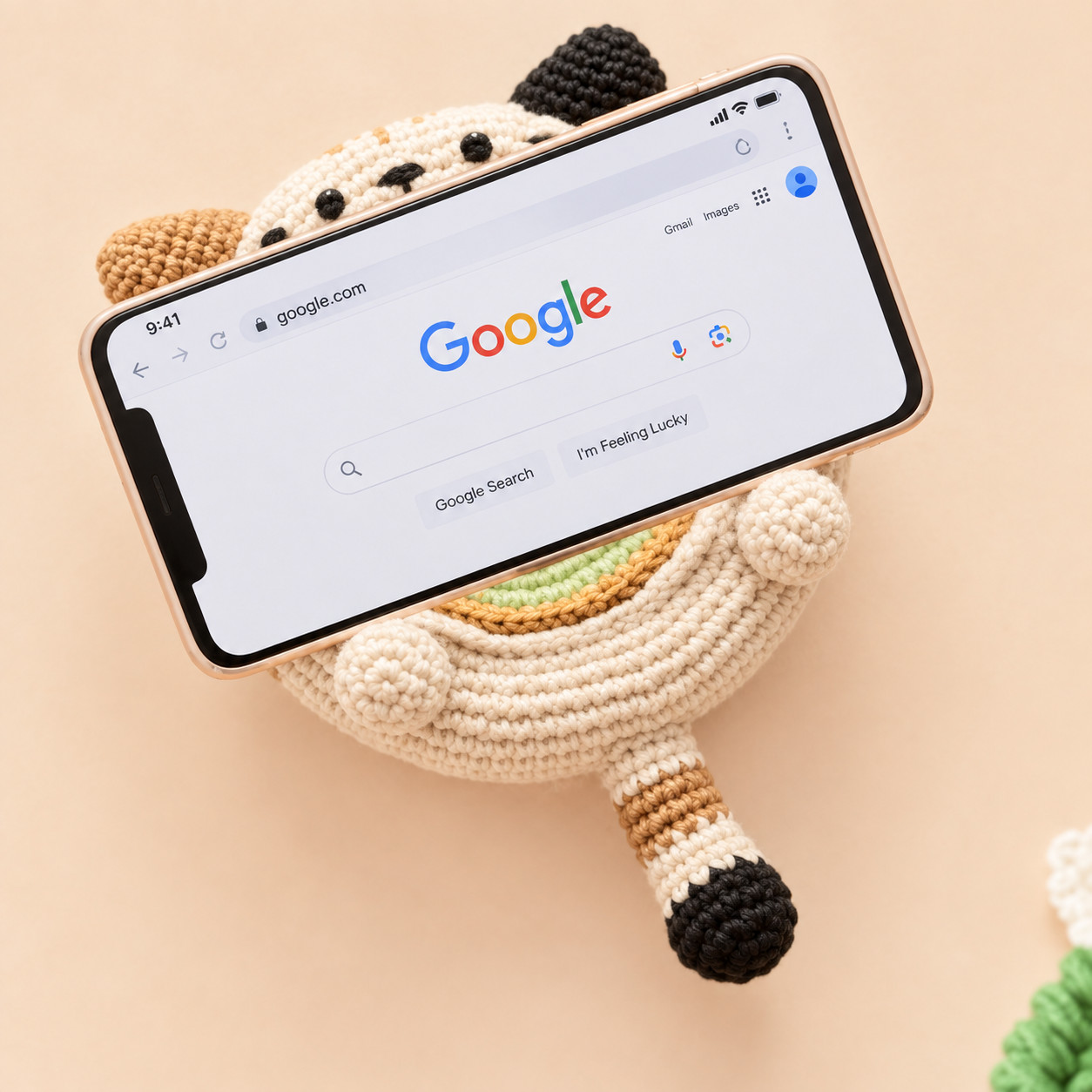 A phone showing a web search page propped sideways against the cream crocheted cat dish on a peach background, the cat's black ear and brown ear showing above it, two round paws holding it from below, and the cat's striped tail with a black tip