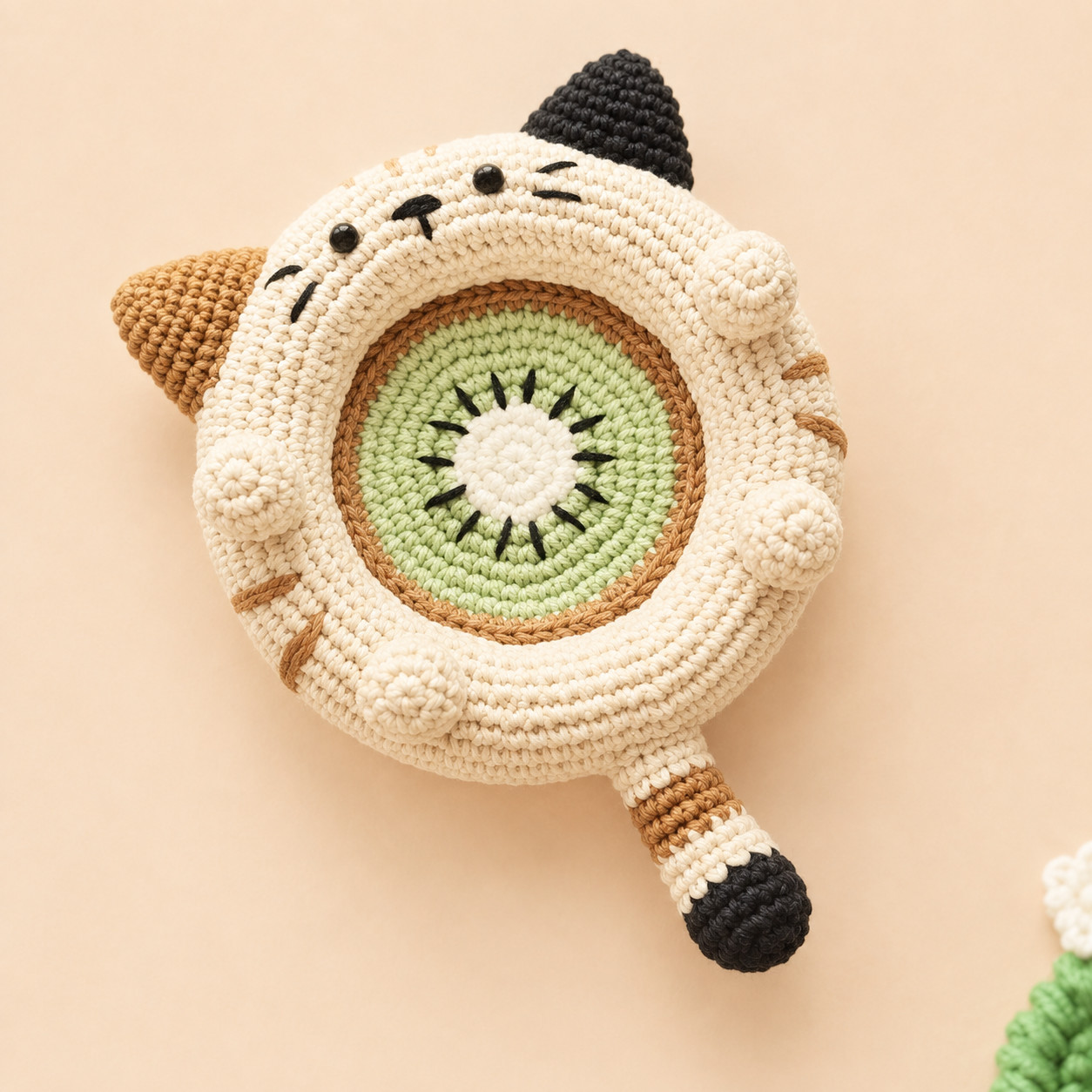 A cream crocheted cat dish seen from above on a peach background, with a kiwi slice in the well: a white centre ringed with black seeds, then green and a brown edge. The cat has one black ear and one brown ear, black eyes and whiskers, brown stripes on the rim, four round paws and a long tail striped cream and brown with a black tip