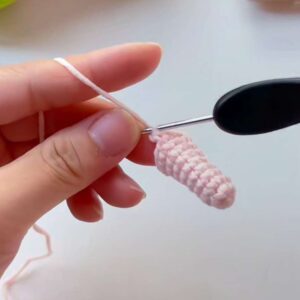 One small pink crocheted arm, a narrow cone, still on the hook between finger and thumb
