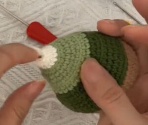 A crocheted tree held on its side in two hands before its leaves go on, the cream tip towards the camera: a band of mint green, then army green, then brown, with a red-handled hook on the white blanket behind