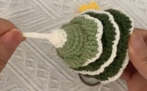 A crocheted Christmas tree held on its side with all three tiers of cream-edged leaves on, a cream cord being drawn out through the top with the fingertips, and a metal ring showing below the bottom edge