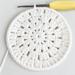 The white openwork centre of a crocheted coaster at the end of its fifth round, rows of small holes running out from the middle, with a yellow-handled hook resting in its last loop