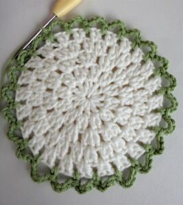 Green chain loops being crocheted round the edge of a white openwork coaster centre, the hook still in the work